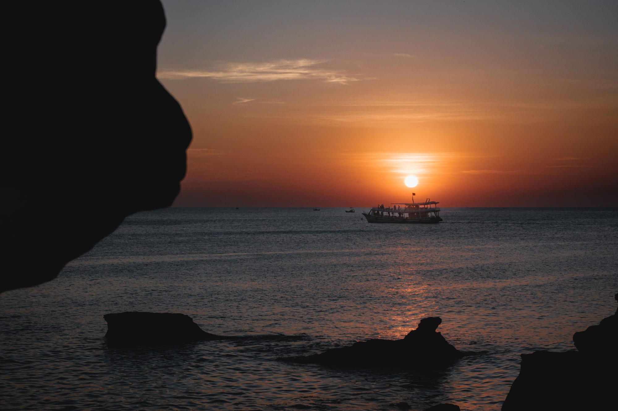 A small fishing boat silhouetted against a glowing orange sunset over the calm sea at Ong Lang Beach, Phú Quốc